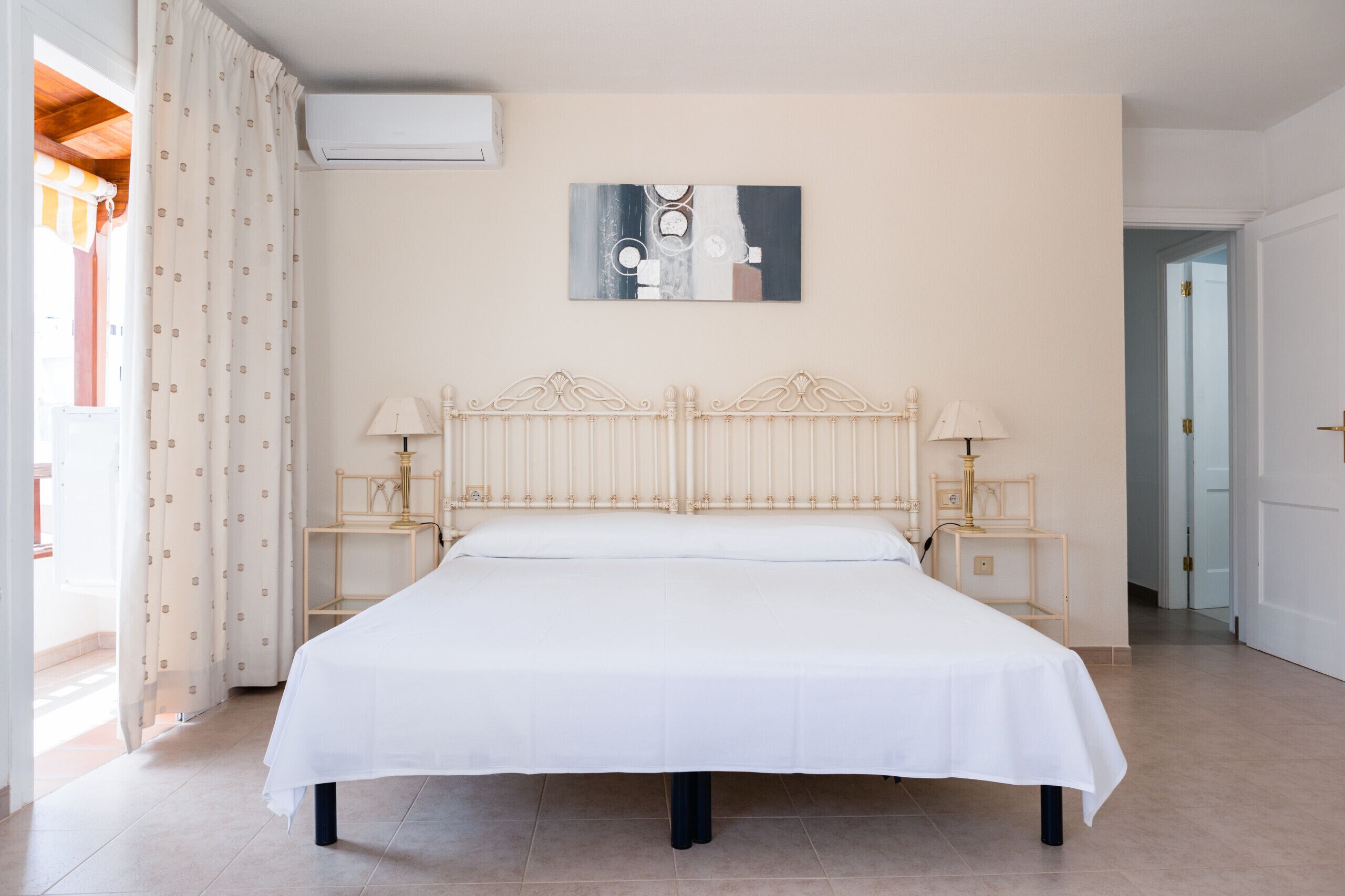 A large bed with a white bedspread, two bedside tables with lamps, a wall-mounted artwork, and a window with curtains.