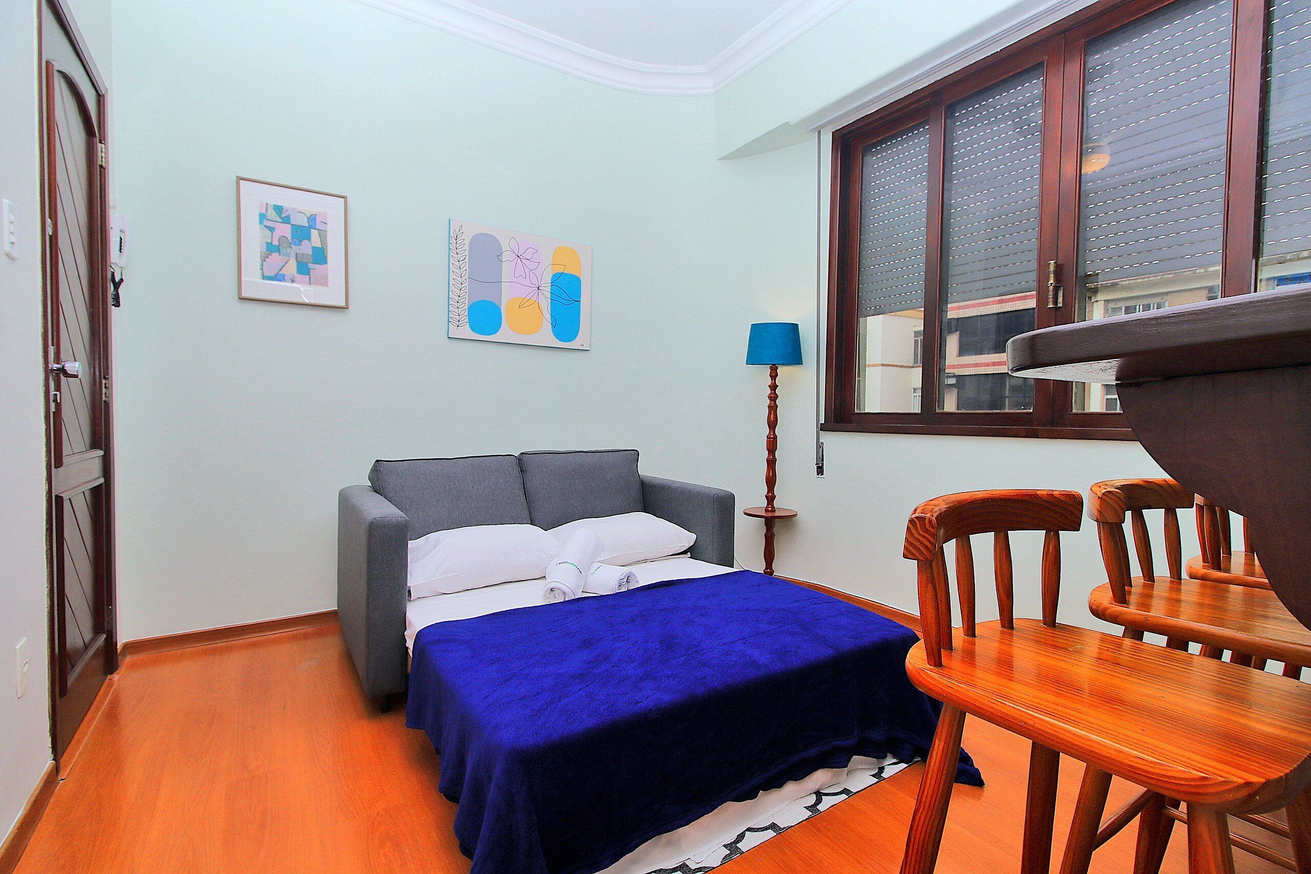 A bedroom with a bed, a grey sofa, a wooden chair, a blue blanket, and a framed abstract art piece on the wall.