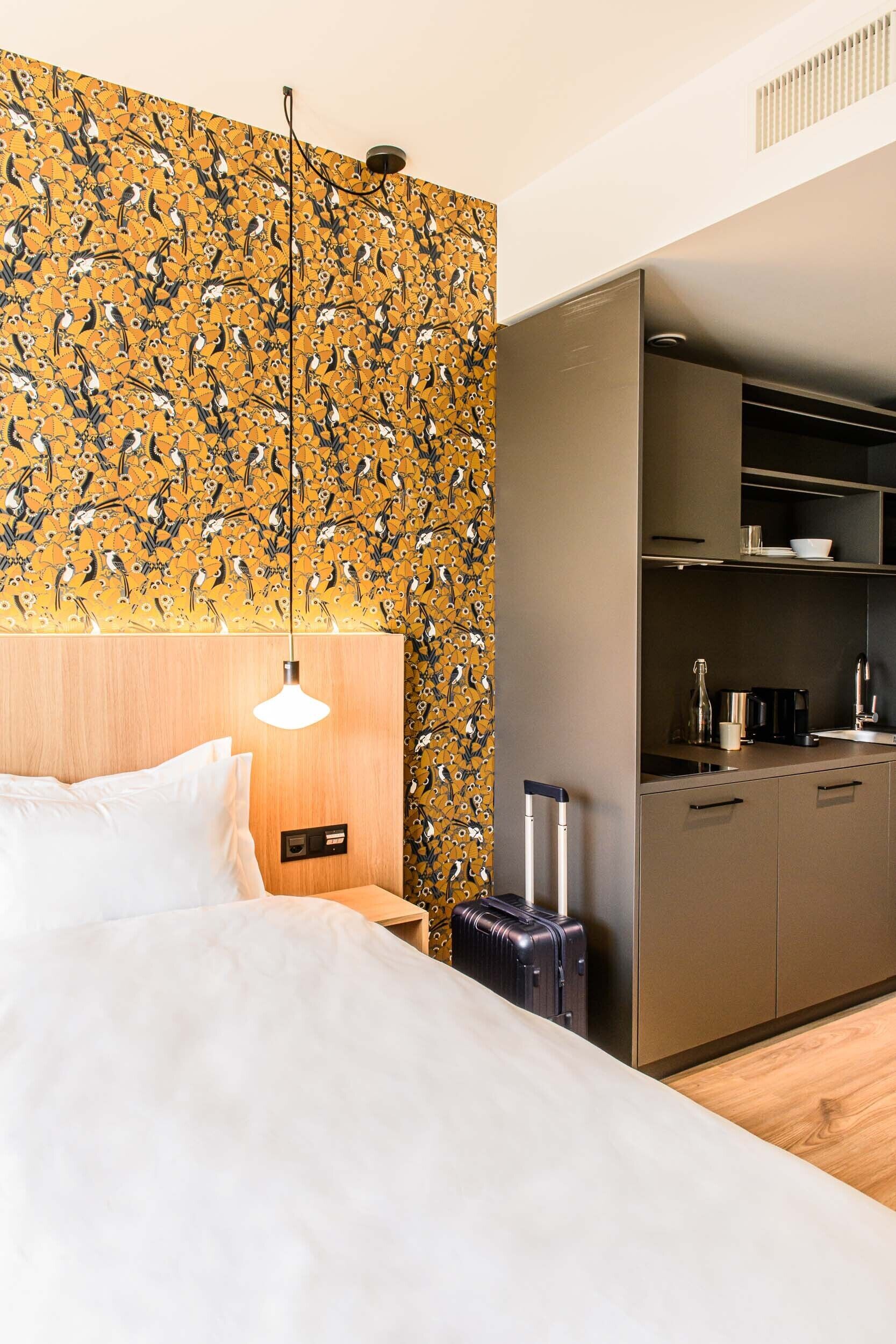 Una habitación de hotel con cama, mesita de noche, maleta y una cocina pequeña con armarios y encimera.