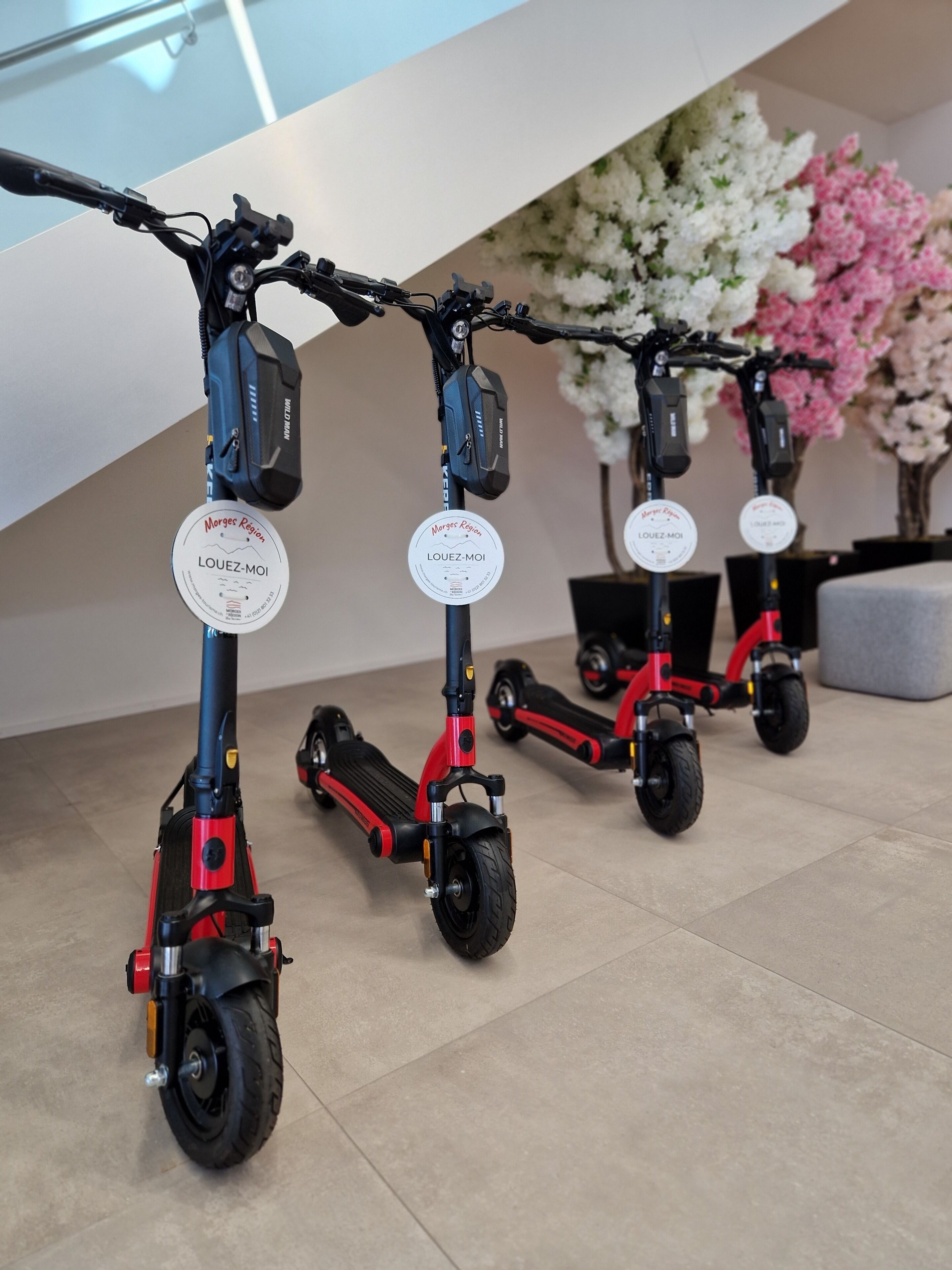 Tre scooter elettrici con adesivi che riportano 'Morges Region', disposti in interni su un pavimento piastrellato, con un'aggiunta floreale sullo sfondo.