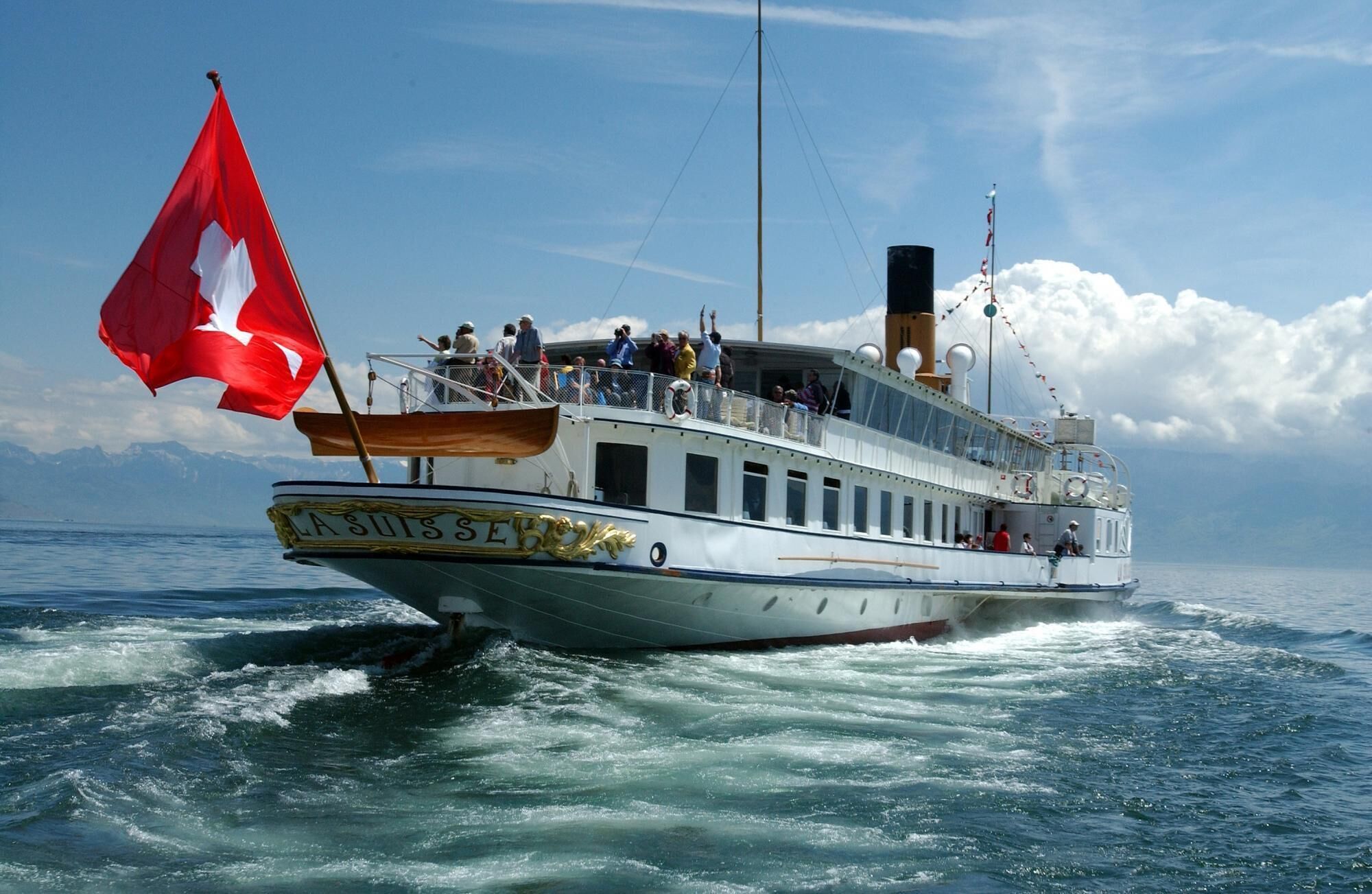 A boat with a white hull and a red flag, named "LA SUISSE," sailing on the water.