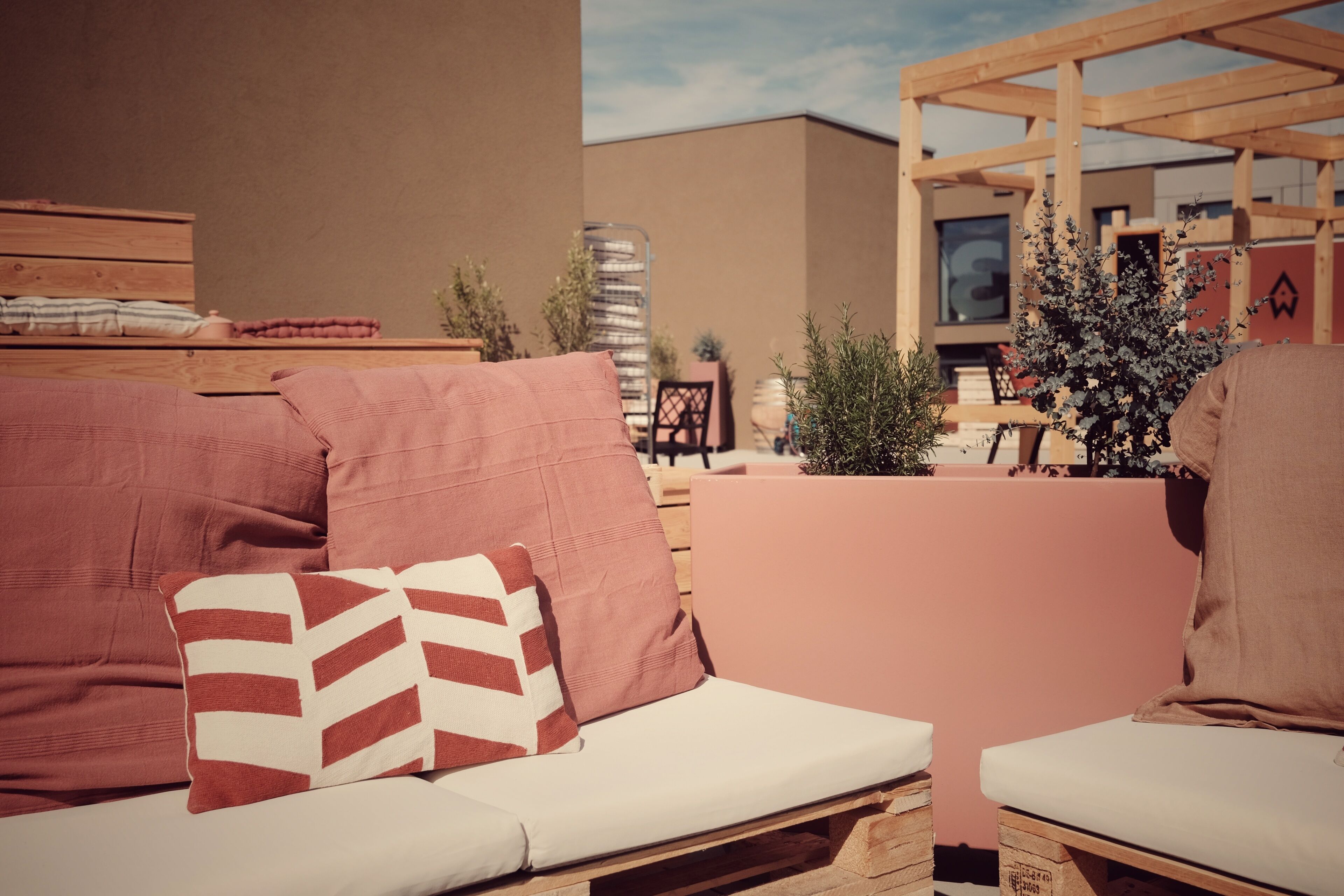A wooden outdoor seating area with cushioned benches and pillows, a small Christmas tree, and a building in the background.