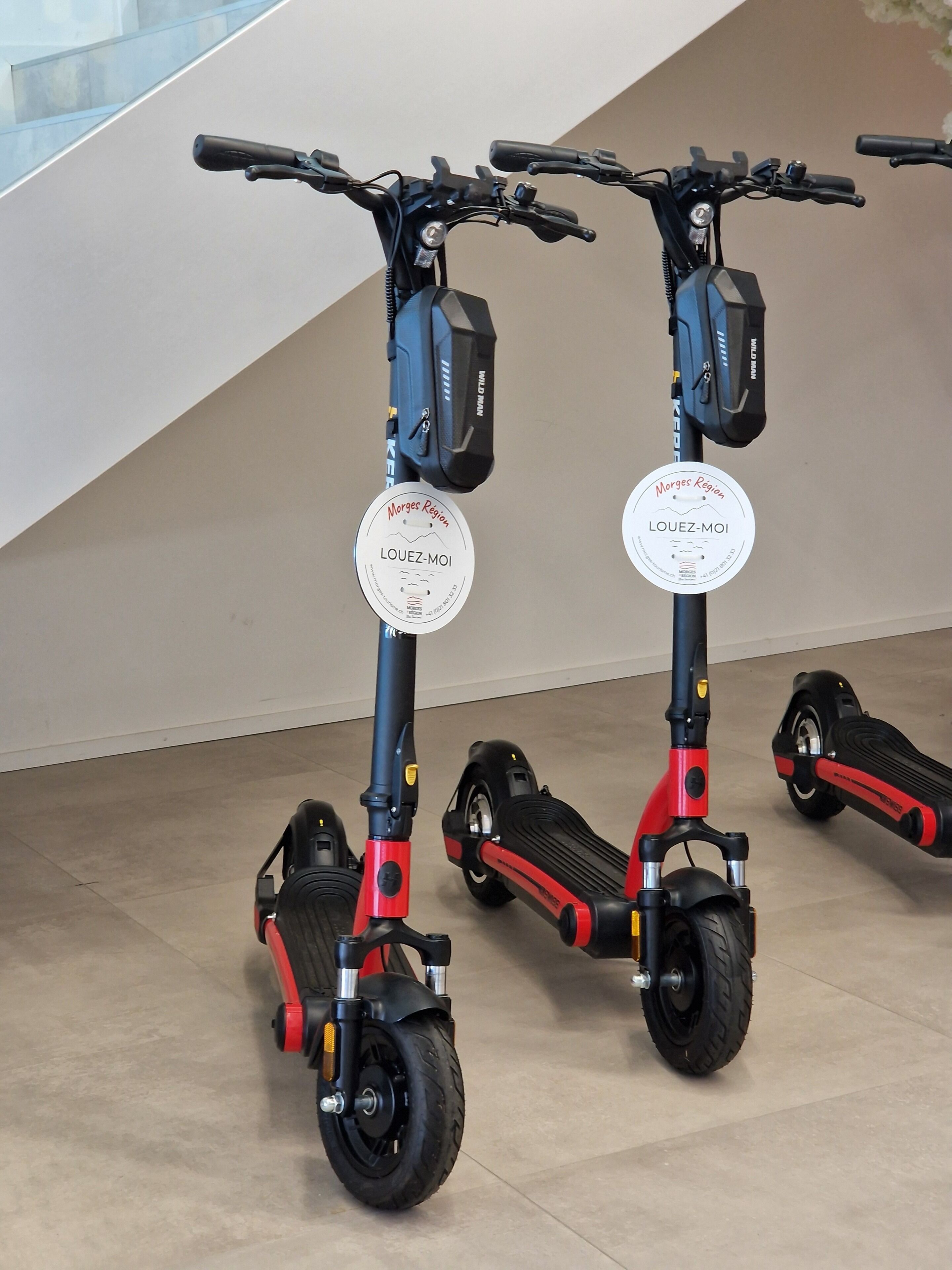 Two electric scooters with 'Morges Region LOUZ-MOI' stickers, placed indoors on a tiled floor.
