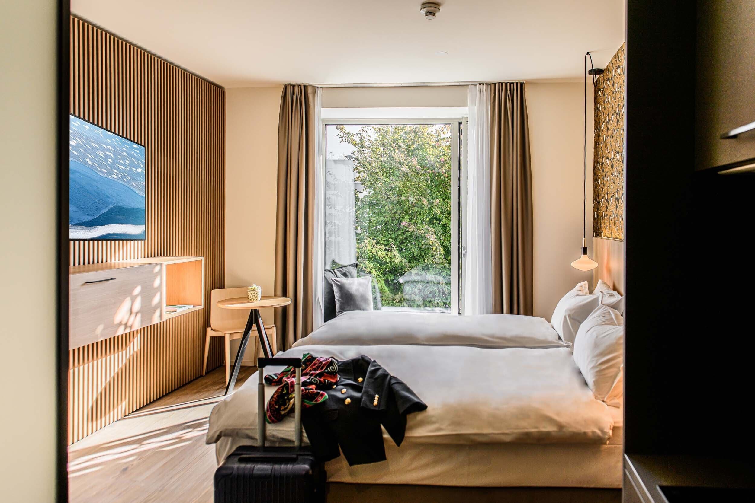 A hotel room with a large bed, a desk, a chair, a TV, and a window with a view of greenery.