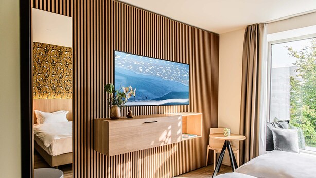 A modern hotel room with a large TV mounted on a wooden panel wall, a bed, a small round table, and a window with a view of greenery. - Afterwork Hotel (Etoy)