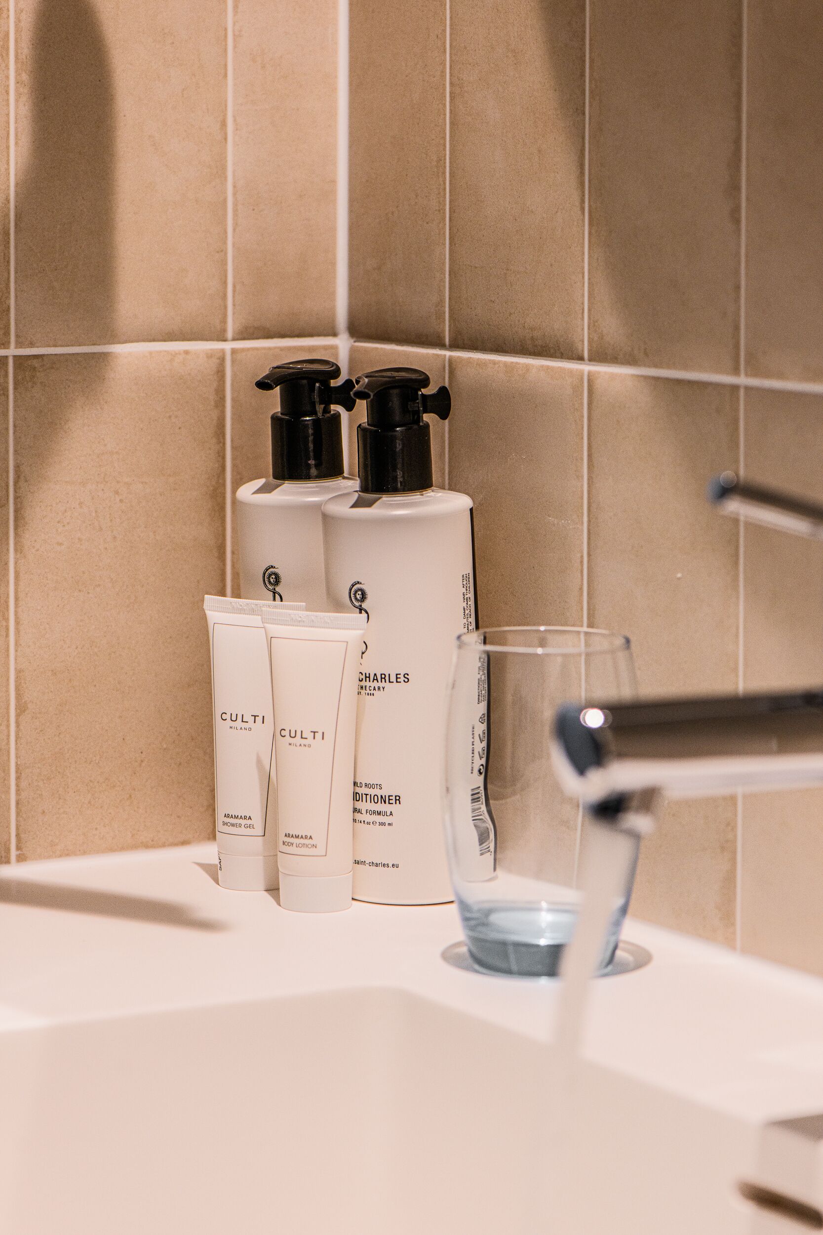 A bathroom with toiletries including a showerhead, a glass of water, and two bottles of Charles & Armandis body lotion.