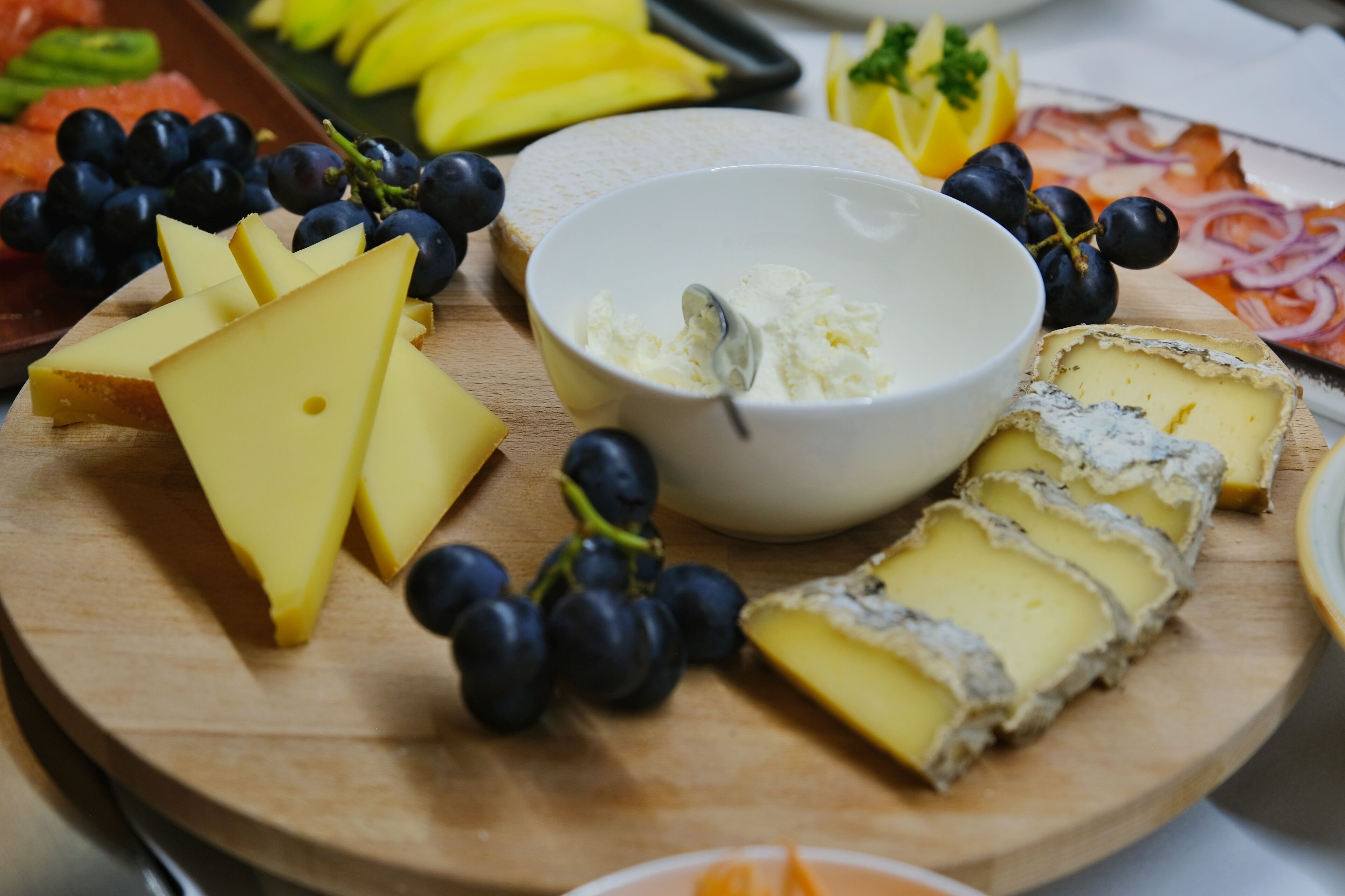 A wooden board with various cheeses, grapes, and a bowl of a creamy spread.