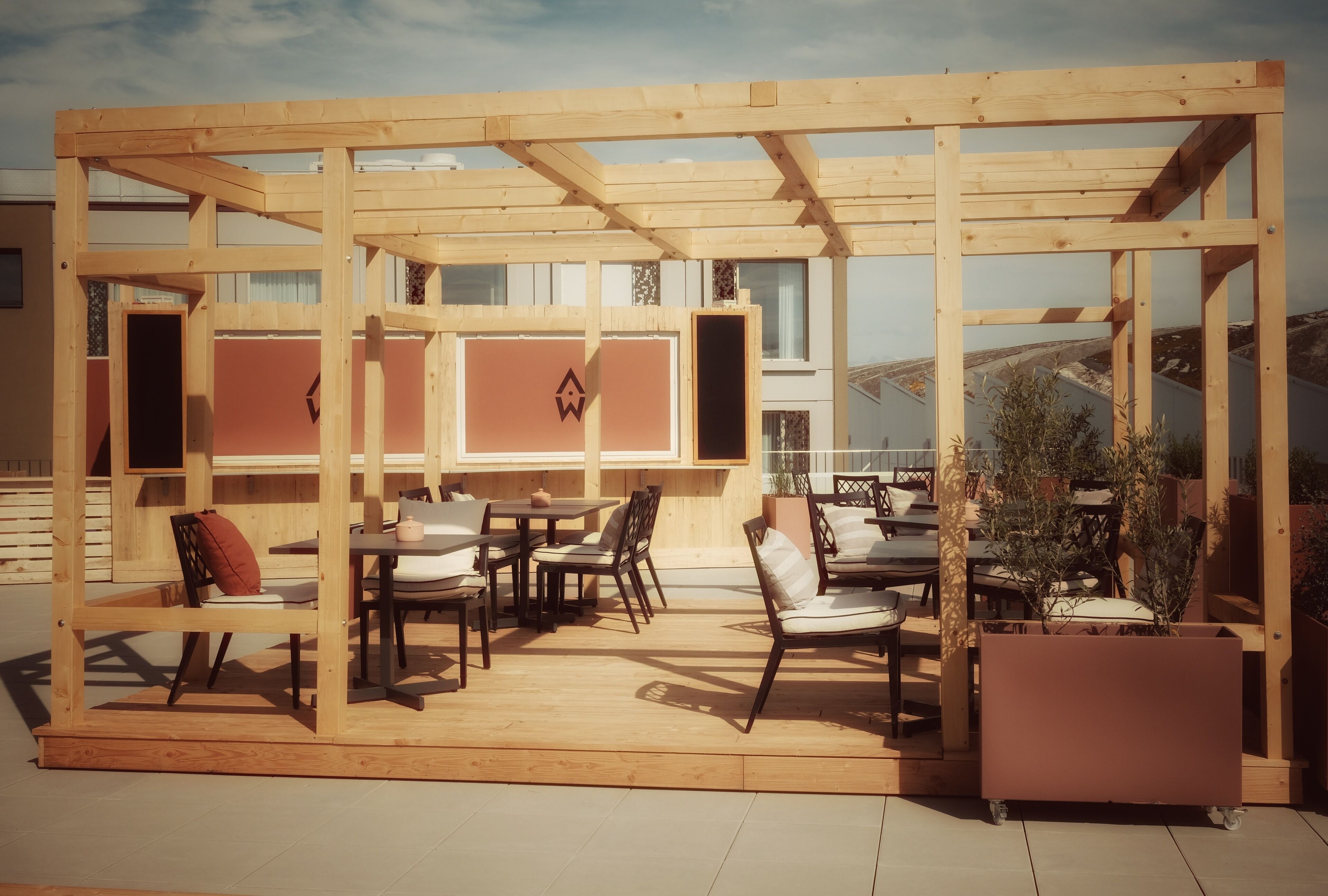 Un espace extérieur aménagé avec des tables et des chaises en bois, délimité par une structure en bois, et agrémenté d’une plante en pot.