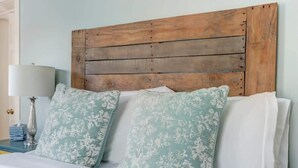 Een bed met houten hoofdbord, twee gestreepte kussens, een wit kussen, een nachtkastje met een lamp en een zakdoos.