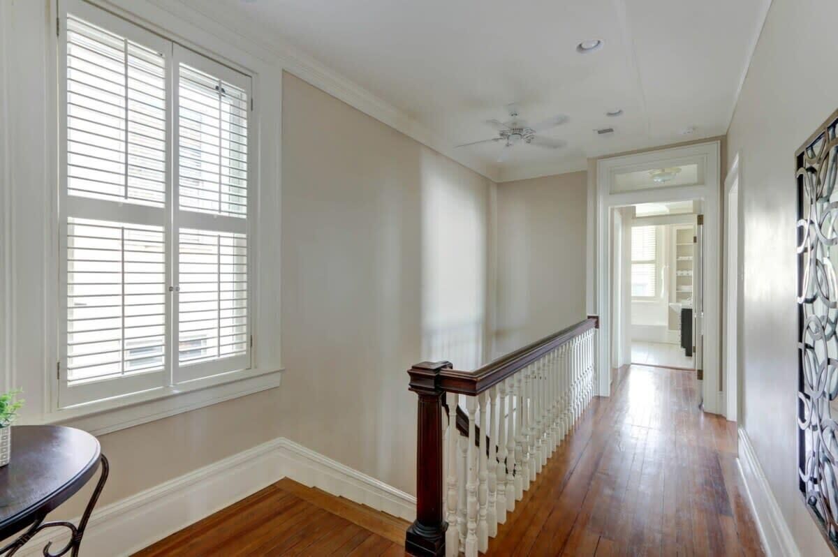 A hallway with wooden flooring, white walls, a ceiling fan, and a staircase with a white railing.