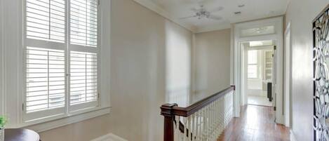 A hallway with wooden flooring, white walls, a ceiling fan, and a staircase with a white railing.