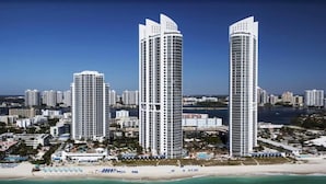 A coastal cityscape with two prominent high-rise buildings, a beach with umbrellas, and a clear sky. - Luxurious Resort|Balcony Bay View|Amazing Pool (Sunny Isles Beach)