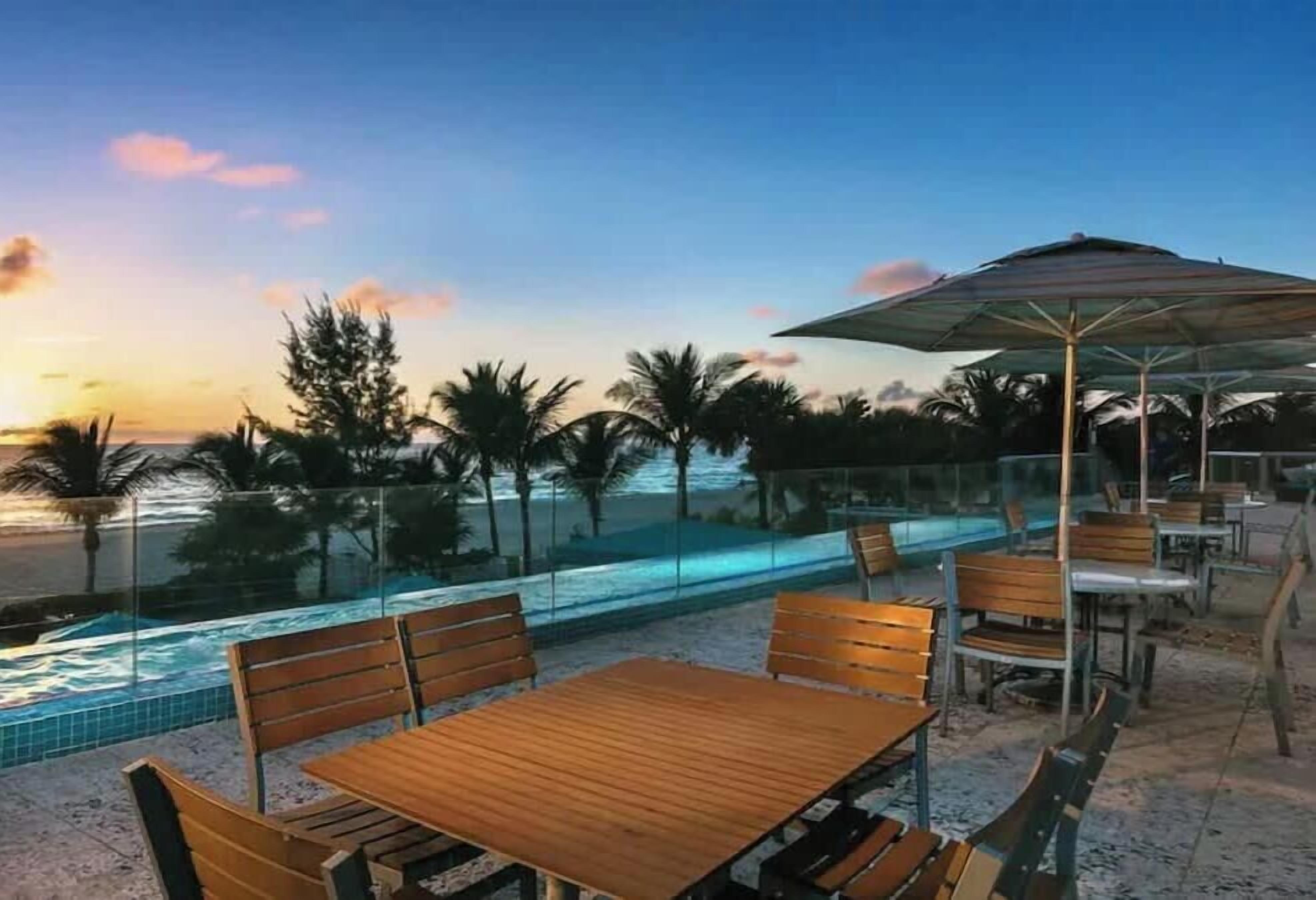 An outdoor seating area with a pool, wooden tables, and chairs, overlooking a body of water.
