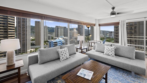 Habitación de hotel moderna con dos sofás grises, una mesa de centro de madera y un ventanal con vistas a la ciudad.