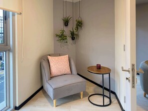 A corner room with a grey armchair, a small round table with a red object, and hanging plants on the wall.