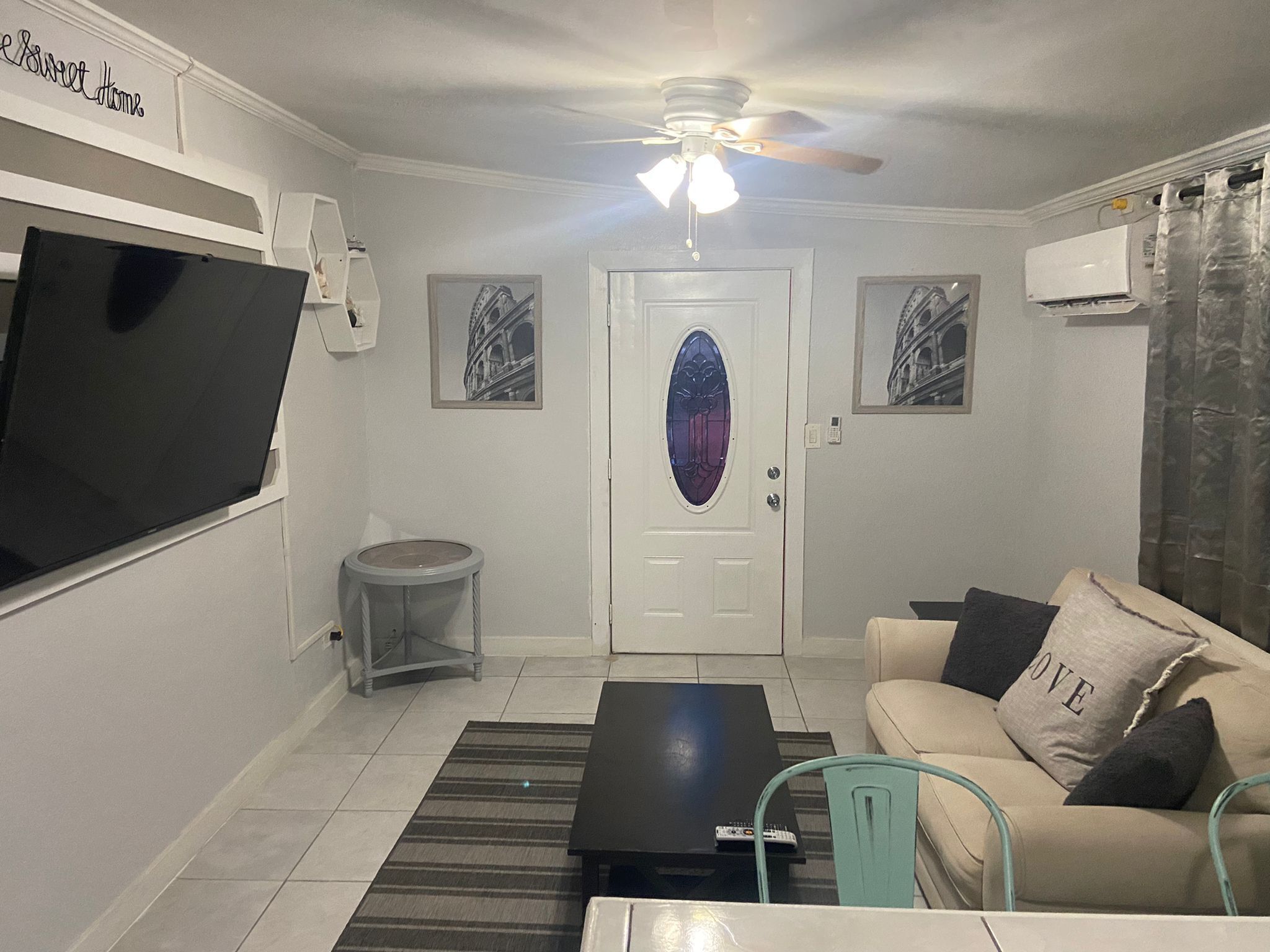 Un salon équipé d’un téléviseur à écran plat, d’un ventilateur de plafond, d’une porte ornée d’un vitrail et d’un canapé agrémenté d’un coussin sur lequel est brodé le mot « AMOUR ».