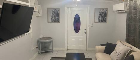 Un salon équipé d’un téléviseur à écran plat, d’un ventilateur de plafond, d’une porte ornée d’un vitrail et d’un canapé agrémenté d’un coussin sur lequel est brodé le mot « AMOUR ».