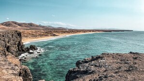 A coastal landscape with rocky cliffs, a sandy beach, and distant mountains under a clear sky. - Ultimate Ocean View El Cotillo (El Cotillo)