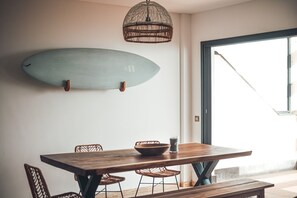 A dining area with a wooden table, wicker chairs, and a surfboard mounted on the wall. - Ultimate Ocean View El Cotillo (El Cotillo)