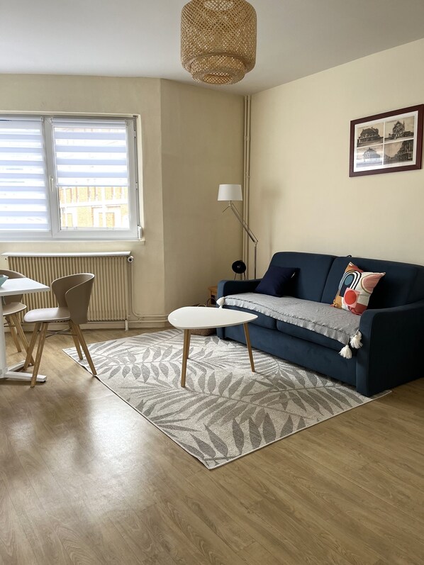A living room with a blue sofa, a white coffee table, a patterned rug, a dining area with chairs, a floor lamp, and a hanging light fixture.