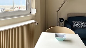 A room with a view of the beach, a radiator, a table with a chair, a lamp, and a sofa with a patterned pillow. - Escale en Duo, studio Plage d'Opale sea view +wifi (Neufchâtel-Hardelot)