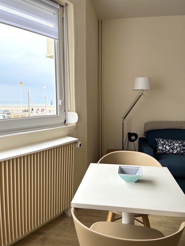 Ein Zimmer mit Blick auf den Strand, ein Heizkörper, ein Tisch mit Stuhl, eine Lampe und ein Sofa mit einem gemusterten Kissen.