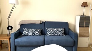 A living room with a blue sofa, two patterned pillows, a white coffee table, a framed picture on the wall, a floor lamp, and a cabinet with drawers. - Escale en Duo, studio Plage d'Opale sea view +wifi (Neufchâtel-Hardelot)