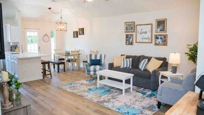 Een ruime woonkamer met een grijze bank, een blauw-witte gestreepte fauteuil en een witte salontafel. Er is een eethoek met een tafel en stoelen, en een keuken met kastjes en een aanrecht.