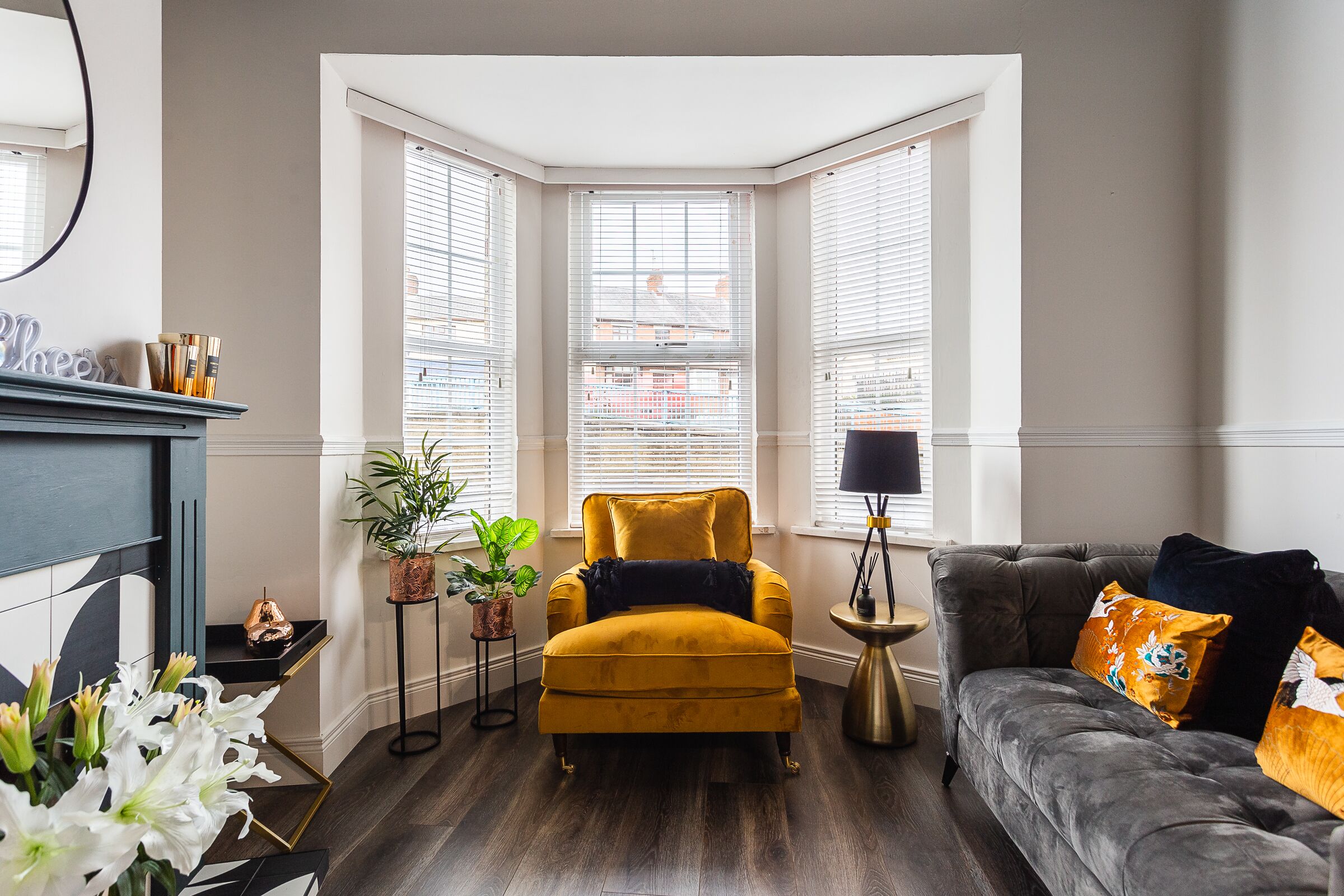 A corner room with a yellow armchair, a grey sofa, and a bay window with blinds.