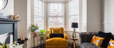 A corner room with a yellow armchair, a grey sofa, and a bay window with blinds.