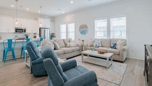 Una sala de estar moderna con un sofá gris, sillas azules, una mesa de centro y una zona de cocina con armarios y electrodomésticos blancos.
