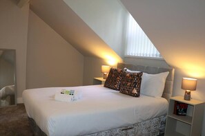 A bedroom with a large bed, two bedside lamps, a bedside table with a book, and a window with blinds.