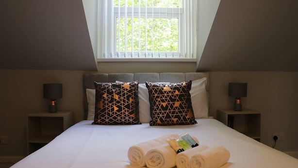 Un dormitorio con una cama grande, dos mesitas de noche, almohadas con estampado geométrico, una ventana con persianas y toallas enrolladas sobre la cama.