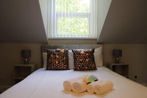 A bedroom with a large bed, two nightstands, geometric patterned pillows, a window with blinds, and rolled towels on the bed.