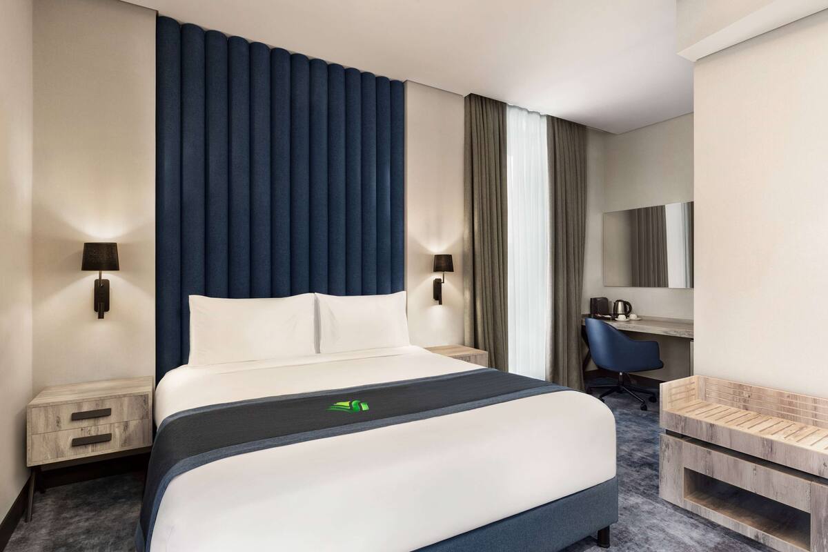 Una camera d'albergo moderna con un letto grande, due comodini con lampade, una scrivania con sedia, uno specchio e una piccola panca di legno.