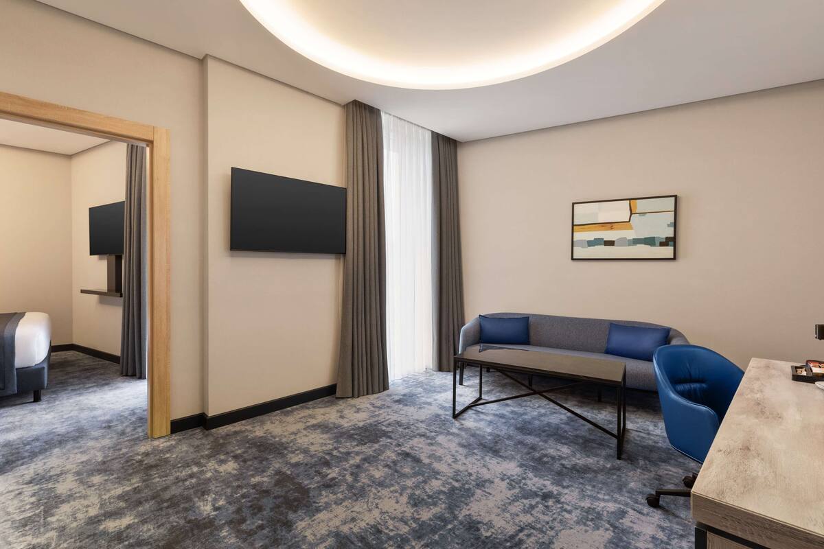Una camera d'albergo moderna con una moquette grigia, un divano grigio, una poltrona blu, un tavolino e un televisore a schermo piatto fissato al muro.