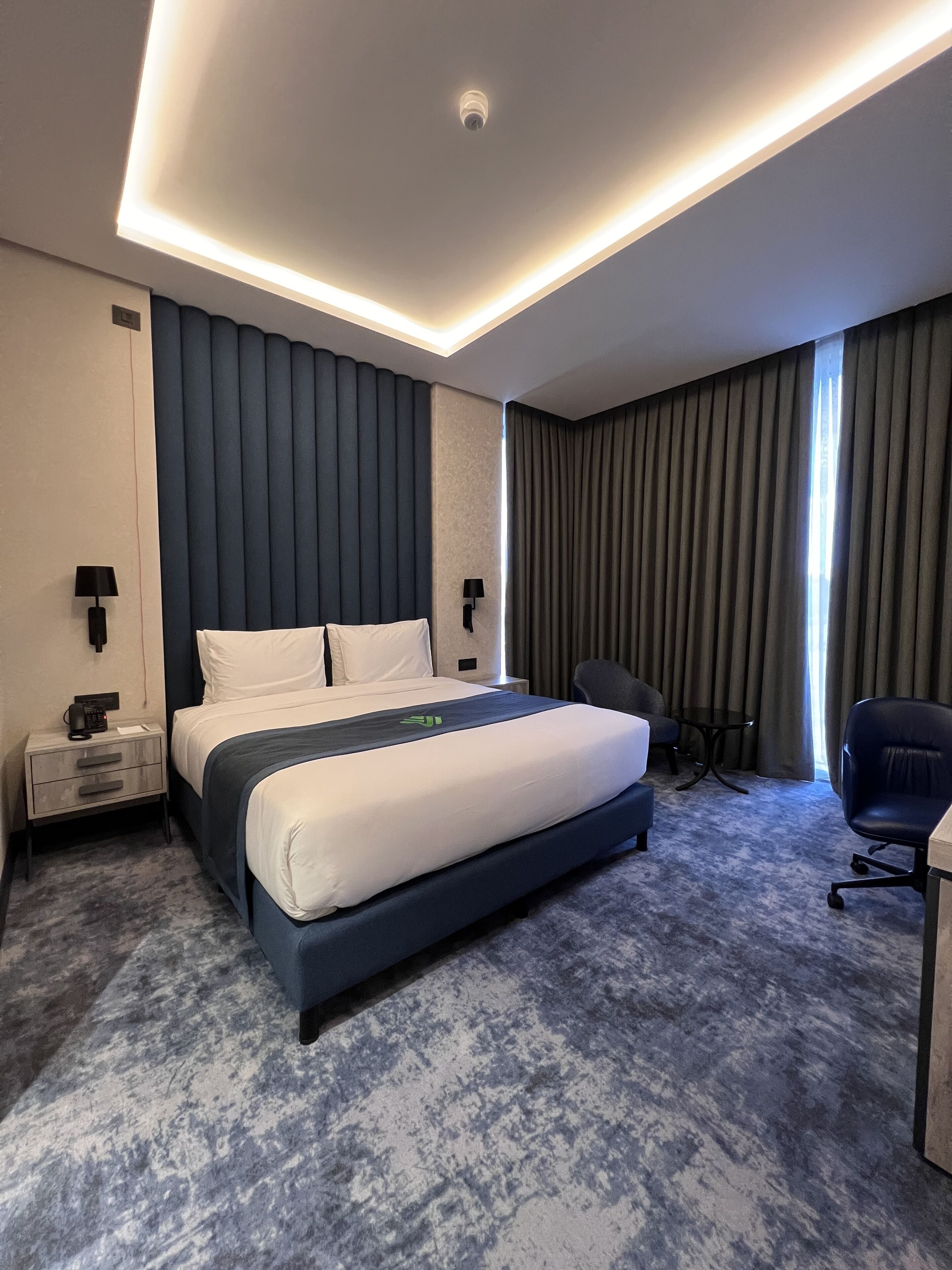 A modern hotel room with a large bed, a desk, a chair, and a view of the city.