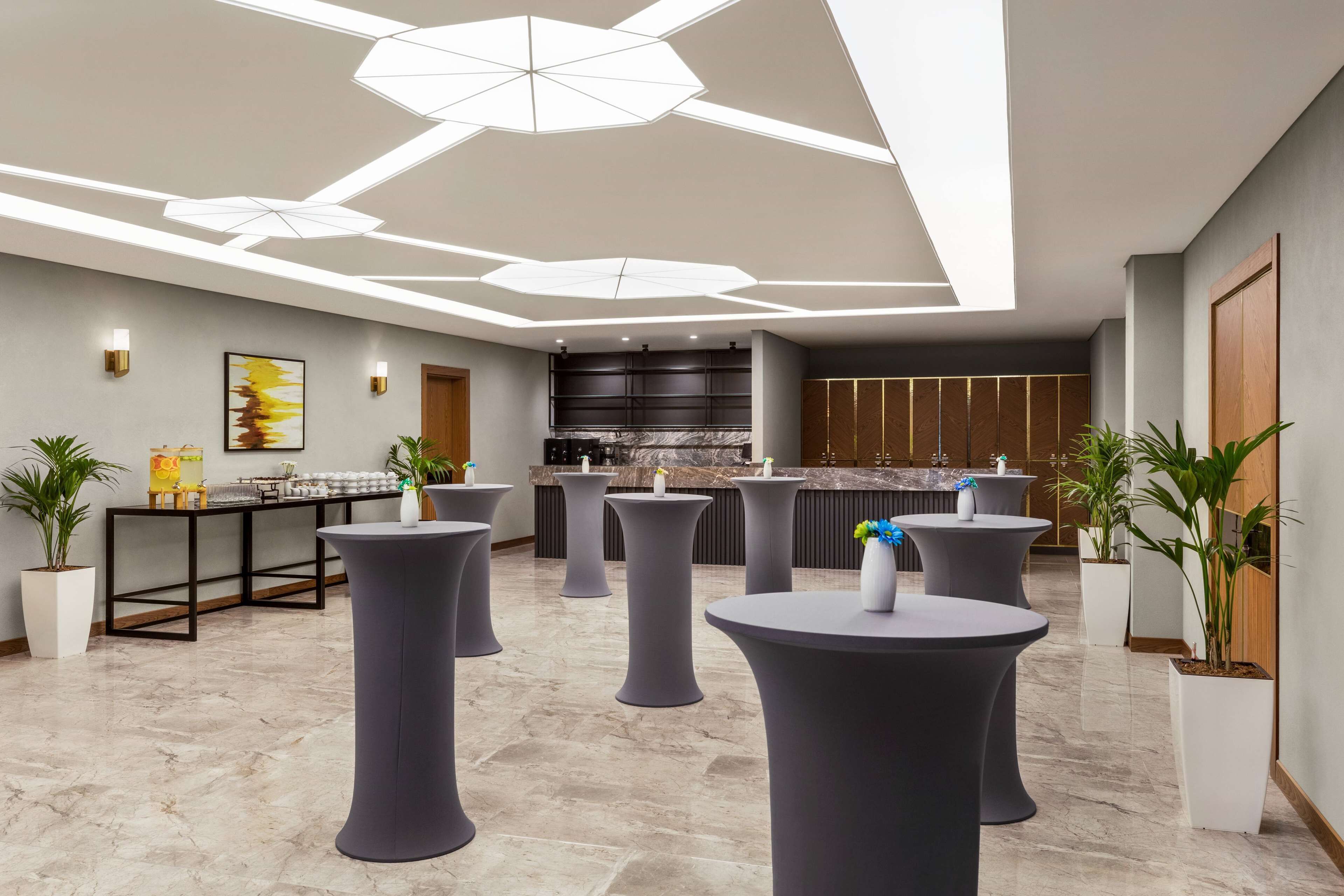 A modern hotel lobby with round tables, potted plants, and a kitchen area in the background.