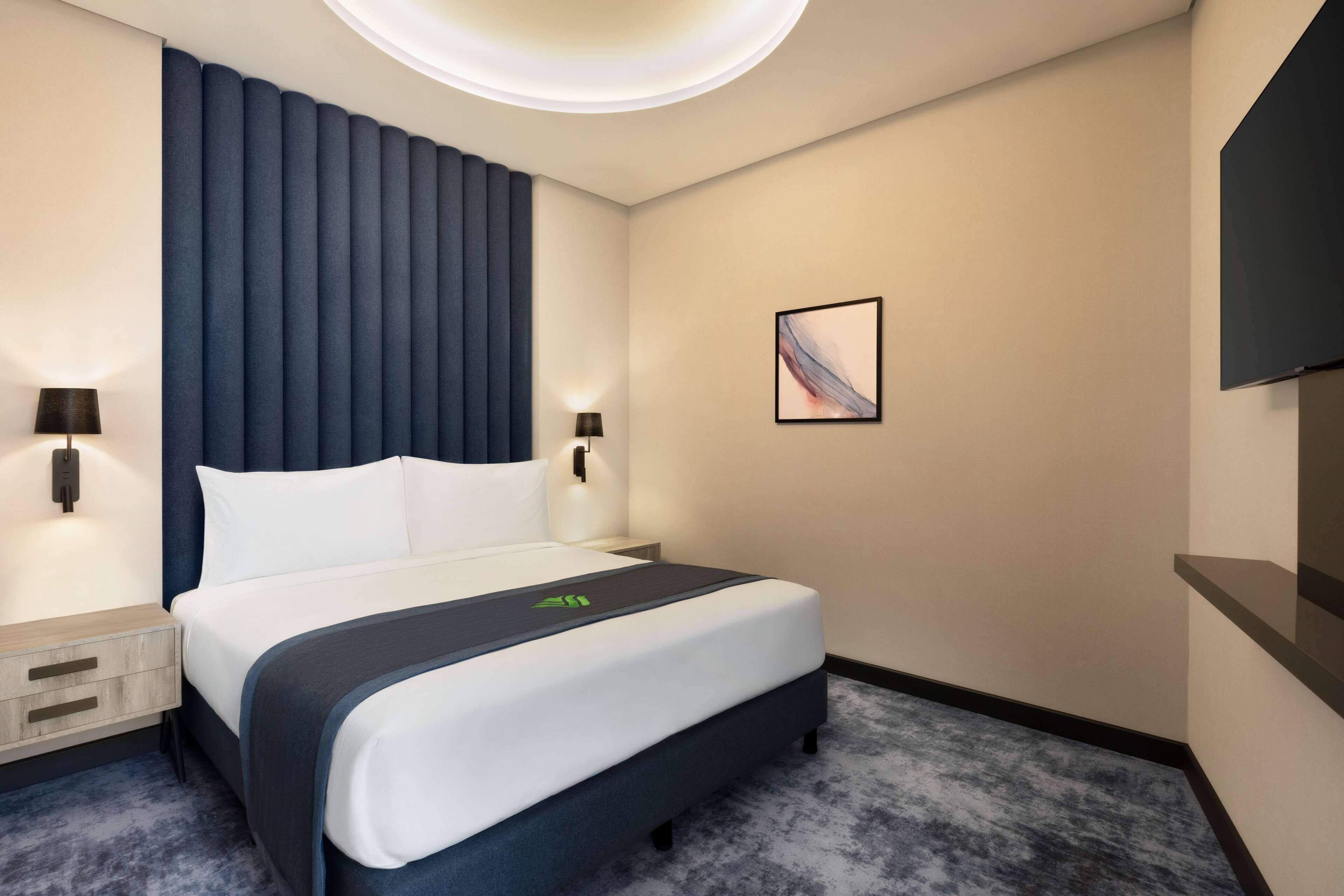 A modern hotel room with a large bed, a headboard with vertical lines, two bedside lamps, a flat-screen TV on the wall, and a small bedside table with a lamp.