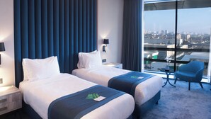 Une chambre d’hôtel moderne avec deux lits, une grande fenêtre donnant sur la ville, un luminaire rond au plafond et une chaise bleue.