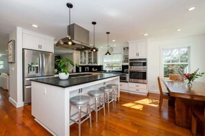 A modern kitchen with stainless steel appliances, a central island with bar stools, and a dining area with a wooden table and chairs. - Relaxing West Cliff Escape with Hot Tub! (Santa Cruz)