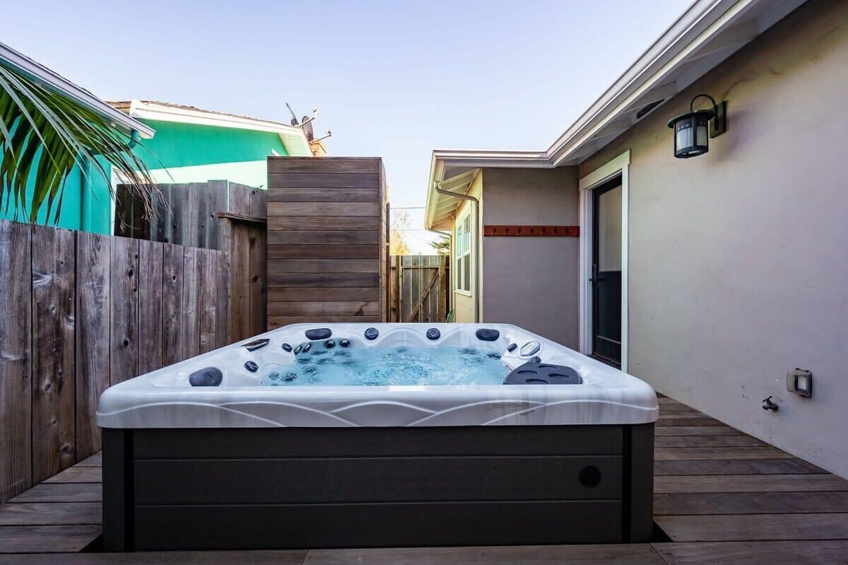 A hot tub in a backyard with a wooden deck, a wooden fence, and a house in the background.