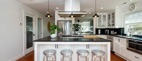 Une cuisine moderne dotée d’un îlot central, d’appareils électroménagers en acier inoxydable, de luminaires suspendus et offrant une vue sur l’extérieur.