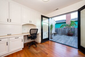 Una habitación con escritorio, silla y puerta corrediza de vidrio que da a un patio exterior con piso de madera y plantas en macetas.