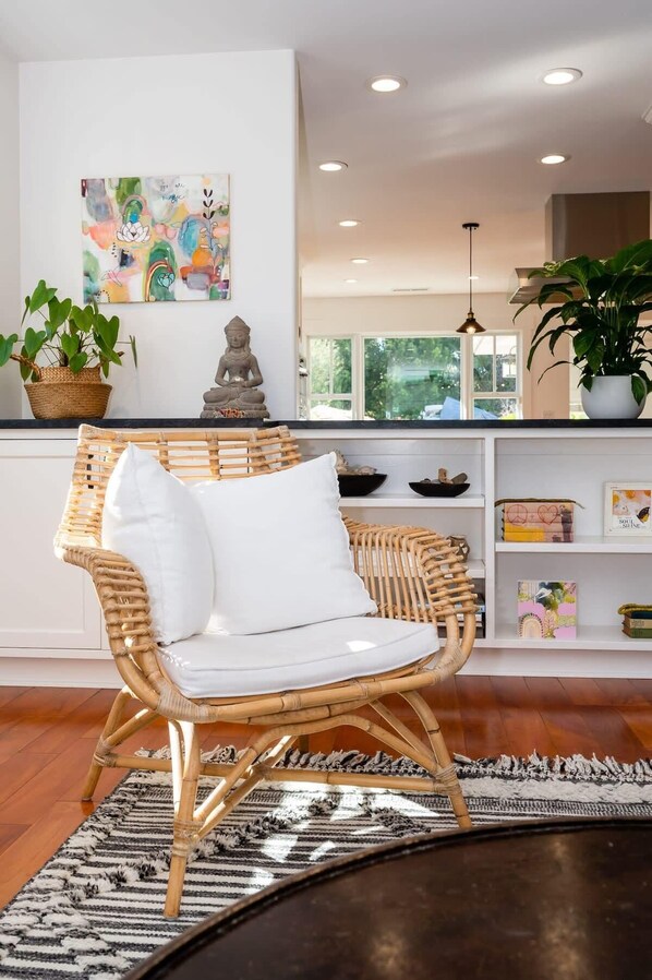 A wicker chair with white cushions in a room with a large window, a painting on the wall, and a shelf with decorative items.