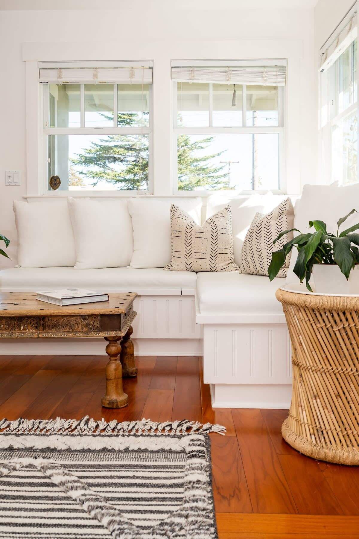 Un sofá blanco con cojines estampados, una mesa de centro de madera y una silla tejida en una habitación luminosa con grandes ventanales.