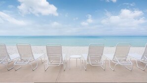 Eine Reihe weißer Liegestühle und ein kleiner Tisch an einem sandigen Strand unter blauem Himmel und ruhiger See.