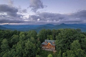 A large, two-story lodge surrounded by dense forest, with a view of distant mountains under a cloudy sky.