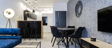 A modern living room with a blue sofa, dining table, and chairs, a coffee table, and a TV on the wall.