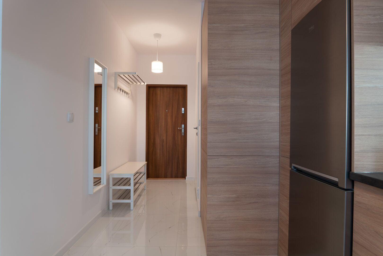 A modern kitchen with a stainless steel refrigerator, a wooden door, and a bench in the hallway.